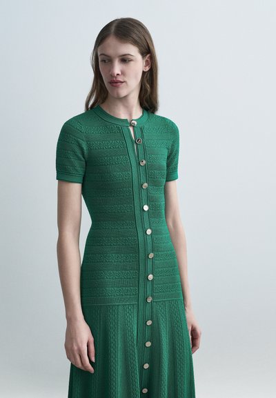 Femme portant une robe verte à manches courtes, boutonnée devant avec des motifs en maille texturée, debout devant un fond uni gris clair.