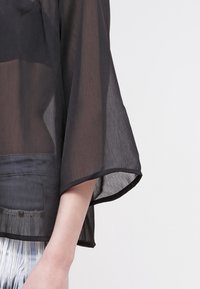 Zwarte doorschijnende top met brede klokmouwen, voorzien van een zachte textuur en een losse pasvorm, vergezeld van een effen band aan de zoom.