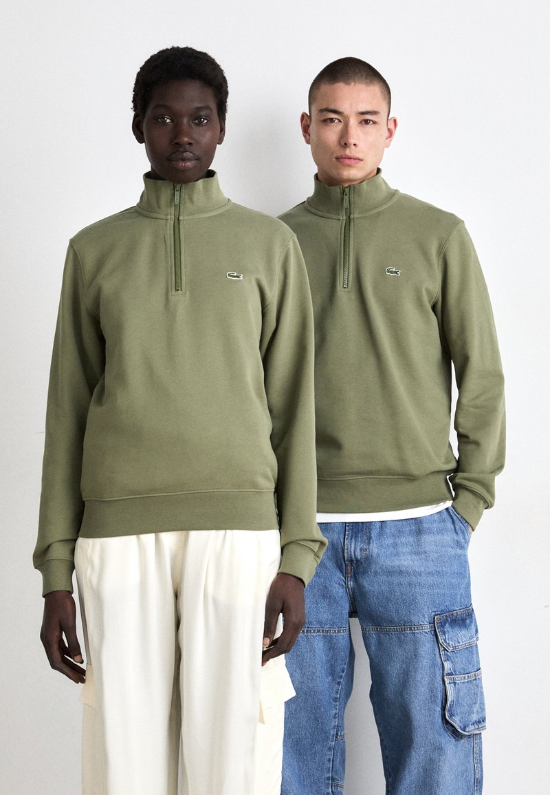 Olivengroene zip-up sweatshirts met hoge kragen en geribbelde manchetten, voorzien van een klein logo op de borst. De modellen worden gestyled met witte en blauwe broeken.