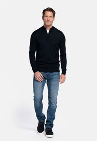 Donker, met rits, pullover sweater, lange mouwen, strak om het lijf. Draagt met lichtblauwe jeans en donkere sneakers. Effen achtergrond.