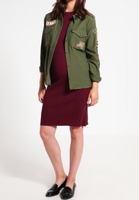 Chemise boutonnée vert olive avec des patchs, superposée à une robe midi côtelée bordeaux. Des mocassins noirs complètent l'ensemble.