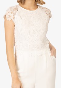 Krajkový crop top v off-white barvě s květinovými vzory, zubatými rukávy a texturovaným povrchem. V kombinaci s krémovými kalhotami.