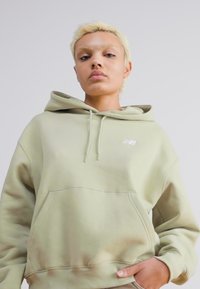 Sweatshirt verde claro com bolso canguru frontal e cordões, apresentando um logotipo branco na área do peito esquerdo e um tecido suave e texturizado.