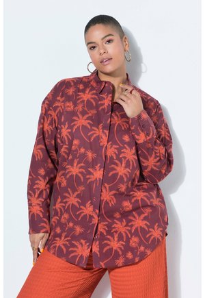 Kvinde med kort hår iført store øreringe, en vinrød skjorte med knapper og orange palmetræsmønster samt teksturerede orange bukser.