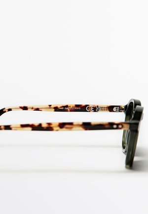 Solbriller med en grøn, rektangulær ramme. Brune skildpaddeskalleremmer med trykt tekst, der inkluderer CE-mærkning og kategorianformation.