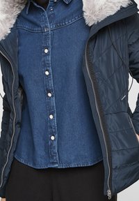 Denimová košile na knoflíky nošená pod nezapnutou námořnickou prošívanou bundou s šedým umělým kožíškem.