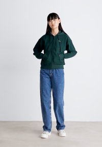 Mørkegrønn hettegenser med glidelås og tætsittende design, kombinert med lyseblå baggy-jeans og hvite joggesko mot en nøytral bakgrunn.