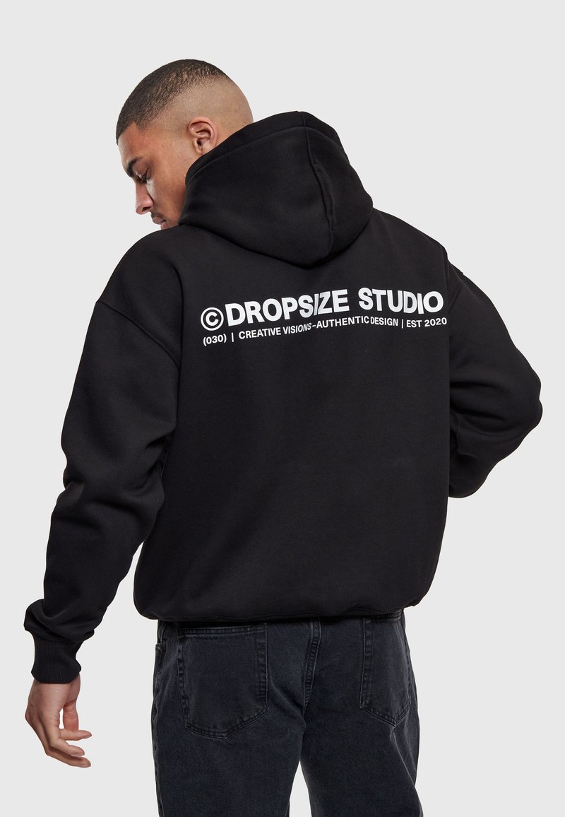Man draagt een zwarte hoodie met kap op, met witte tekst "Dropsize Studio" en slogan op de achterkant, gecombineerd met donkere jeans, tegen een grijze achtergrond.
