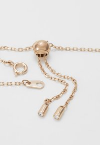 Colar de corrente em tom dourado com um fecho de bola redonda, com duas extremidades cilíndricas adornadas com cristais transparentes. Comprimento ajustável com correntes interligadas.