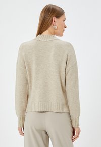 Beige neulepaita, jossa on ribbestyttävä helmaviimeistely ja hihansuut, varustettu korkealla kaula-aukolla ja rentolla mitoituksella, valmistettu pehmeästä, teksturoidusta kankaasta.