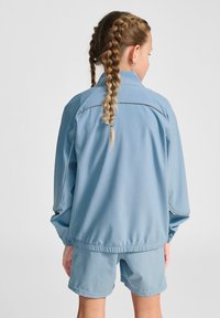 Ljust blå idrottsjacka med elastiska ärmslut, ståkrage och matchande shorts. Slen yta med subtila sömmar och utan mönster.