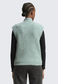Gilet en tricot sans manches de couleur vert menthe, avec une texture côtelée. Col montant haut, coupe légèrement oversize, porté par-dessus un haut à manches longues noir.