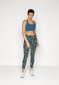 Sportbh i teal med tykke stropper, kombinert med tettsittende leggings med et blått og grønt blomstermønster og hvite joggesko.