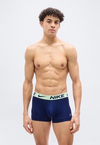 Νεαρός άνδρας με σγουρά μαλλιά, στέκεται μπροστά σε λευκό φόντο, φοράει μπλε σκούρα εσώρουχα μπόξερ Nike με λωρίδα μέσης σε ανοιχτό πράσινο χρώμα.