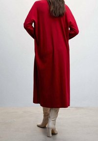 Personne portant un long manteau rouge, les mains dans les poches, chaussée de bottes beige montant jusqu'aux genoux, debout sur un sol gris devant un mur clair uni.