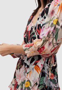 Robe florale avec une base noire ornée de motifs floraux colorés, un décolleté en V profond, des manches fluides avec des poignets froncés et un détail de ceinture à nouer.