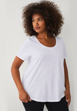 Vrouw met krullend haar, draagt een wit T-shirt met korte mouwen en ronde hals en een zwarte broek, kijkt naar links tegen een effen achtergrond.