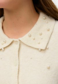 Krēmkrāsas adīts kardigans ar apkakli, dekorēts ar apaļām pērlēm un centrālu pogu aizdari. Mīksta tekstūra.