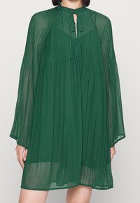 Zielona, plisowana sukienka z szyfonu z wysokim dekoltem, otworem w kształcie klucza oraz długimi, zwiewnymi rękawami. Lekka i przewiewna tekstura.