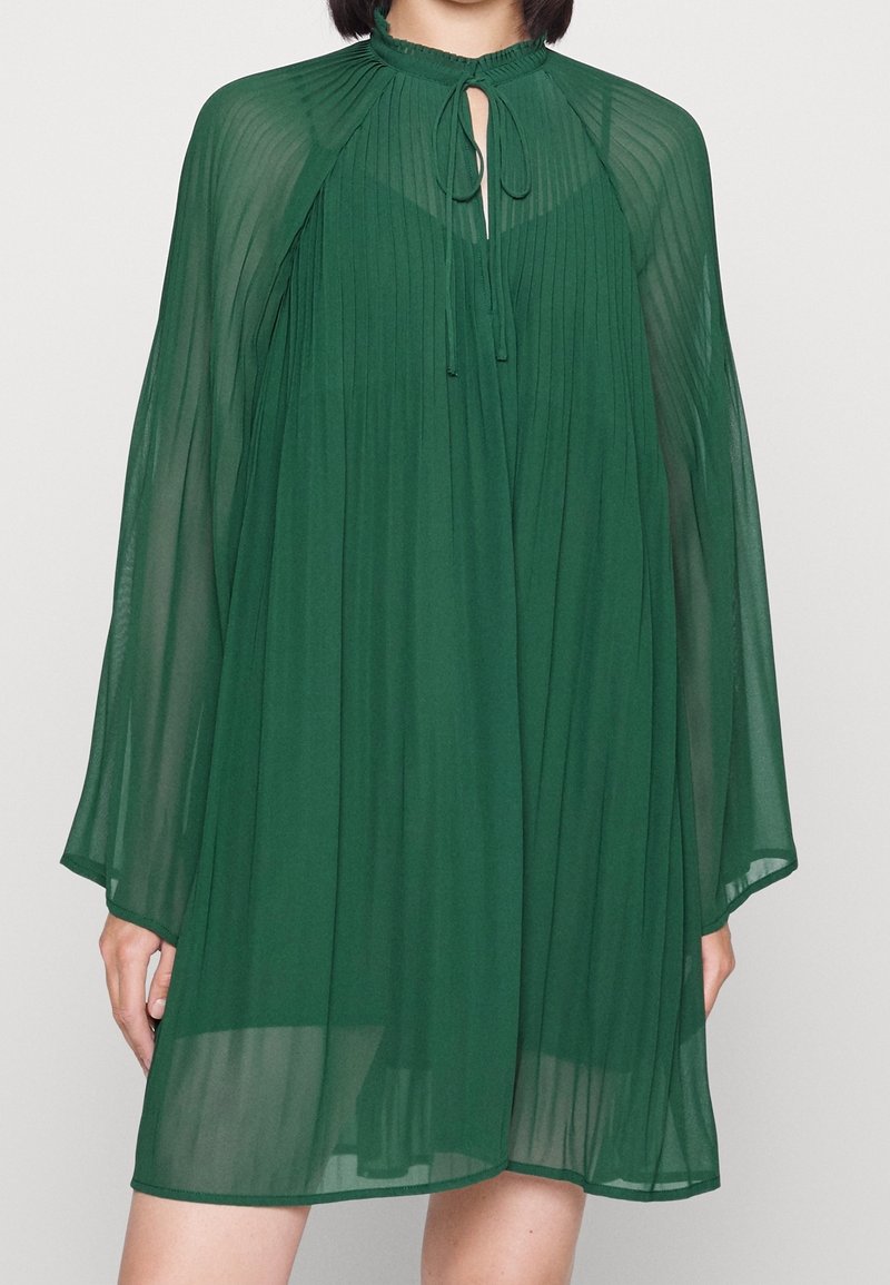 Zielona, plisowana sukienka z szyfonu z wysokim dekoltem, otworem w kształcie klucza oraz długimi, zwiewnymi rękawami. Lekka i przewiewna tekstura.