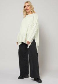 Jasnozielony sweter z dzianiny z bocznymi frędzlami, noszony z czarnymi spodniami o szerokich nogawkach i czarnymi botkami za kostkę. Miękka tekstura, casualowy design.