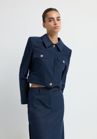 Námořnická zkrácená bunda s velkým límcem a stříbrnými knoflíky, obsahující dvě náprsní kapsy a strukturovaný design. Hladká, texturovaná tkanina.