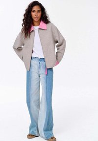 Světle šedá bunda na zip s růžovým límcem, nošená přes bílou košili. Světle modré zvonové džíny s výraznými švy a volným střihem.