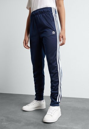 Persoon die marineblauwe Adidas trainingsbroek met witte strepen en witte sneakers draagt, staat op een grijze vloer tegen een lichtgrijze muur.