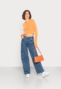 Top laranja cropped de manga longa com fecho de botão, combinado com jeans azul de perna larga. Ténis brancos e uma bolsa laranja completam o look.