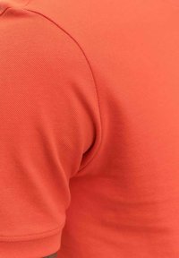 Orange polo-tröja i en strukturväv, med rundad krage och korta ärmar. Sömmar är synliga längs axlarna.