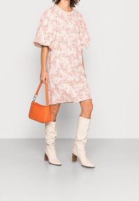 Nainen, joka on pukeutunut löysään valkoiseen ja punaiseen kukalliseen mekkoon, pitää kädessään oranssia käsilaukkua, jalassaan on polveen saakka ulottuvat valkoiset saappaat harmaalla lattialla.