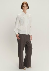 Blusa blanca con lazo, abotonada, de manga larga; combinada con pantalones de raya diplomática gris oscuro. Los zapatos son de punta afilada, color crema.