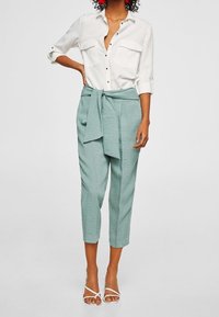 Chemise blanche avec deux poches poitrine et boutons noirs ; associée à un pantalon vert clair avec un nœud à la taille ; sandales blanches ouvertes.