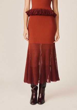 Vestido de punto acanalado color naranja óxido con cintura peplum y panel de falda plisada y translúcida, combinado con botas altas hasta la rodilla de color marrón oscuro.