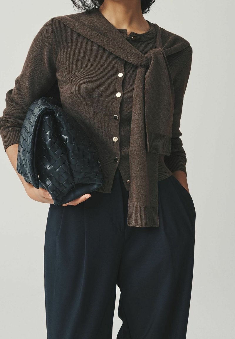 Personne en cardigan marron boutonné avec pull assorti noué autour des épaules, pantalon large noir, tenant une grande pochette tissée noire.