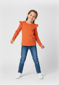 Pomarańczowa koszula z długim rękawem z falbankami na ramionach, połączona z niebieskimi dżinsami i białymi tenisówkami. Prosty fason i casualowy krój.