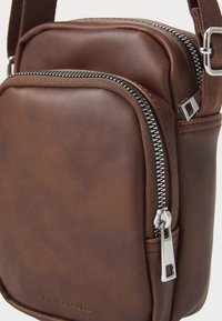Brown leather crossbody bag with silver metal zipper detail, smooth texture, rectangular shape, and adjustable strap. Minimal branding visible.