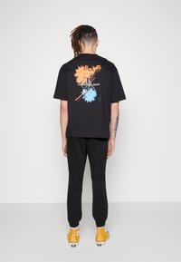 Schwarzes T-Shirt mit buntem Grafikdesign in Orange und Blau auf dem Rücken, kombiniert mit schwarzen Jogginghosen und gelben Turnschuhen.