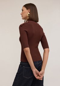 Pulover maro cu maneci scurte, cu dungi, având butoane pe umeri și un design ajustat. Este purtat cu blugi negri، cu buline. Nu sunt prezente alte detalii.