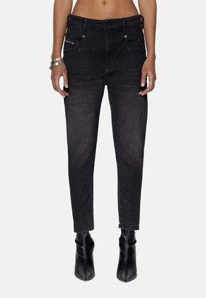 Femme portant un jean noir taille haute en denim légèrement usé, associé à des bottines noires à bout pointu et des bracelets argentés au poignet gauche.