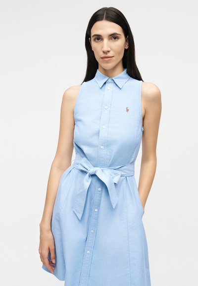 Sieviete ar gariem tumšiem matiem, valkājot bezroku gaisīgas zilas pogām aizdares kleitu, sasietu pie jostas, stāv pret vienkrāsainu gaišu fonu.