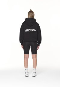 Svart huvtröja med vit text på ryggen, tillsammans med svarta cykelshorts och vita sneakers med svarta detaljer.