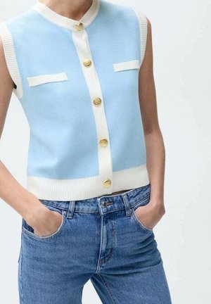 Mouwloze lichtblauwe gebreide gilet met witte bies en gouden knopen, gedragen met blauwe spijkerbroek, handen in de zakken.