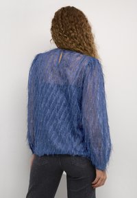 Blå texturerad blus med långa, vida ärmar; genomskinligt material med metalliska trådar och en nyckelhålsdetalj i ryggen. Matchad med mörka jeans.