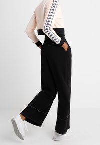Persona con pantaloni neri a gamba larga e sneakers bianche, con le mani nelle tasche, mostrata di lato su uno sfondo bianco.