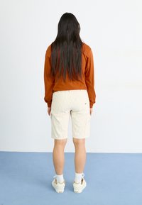 Langermet rustfarget topp og hvite stripete cargo-shorts med lommer. Kombinert med hvite sokker og beige sneakers.