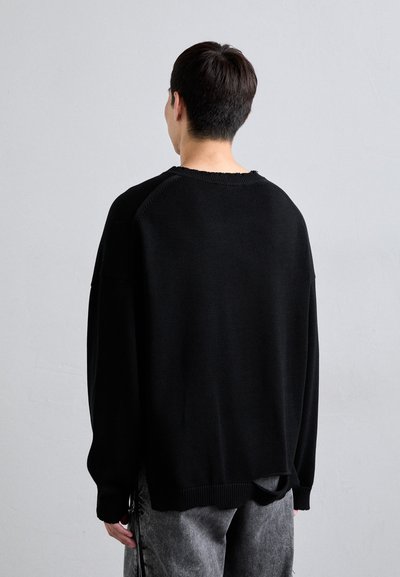 Pull en tricot noir avec une coupe ample, ourlet et poignets côtelés, présentant une finition brute au niveau du col, porté sur un pantalon texturé gris.