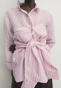 Personne portant une chemise blanche à rayures roses verticales, avec une chemise rayée supplémentaire nouée autour de la taille.