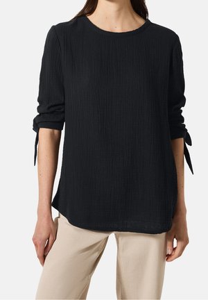 Femme portant un chemisier noir texturé à manches longues avec poignets noués et pantalon beige, visible du cou aux cuisses supérieures.