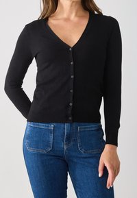 Černý, přiléhavý cardigan s výstřihem do V, zapínáním na knoflíky a dlouhými rukávy; hladká textura; v kombinaci s modrými džíny.